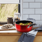 A metallic red nonstick dutch oven pot filled with cooked food on a wooden kitchen counter next to a loaf of bread.