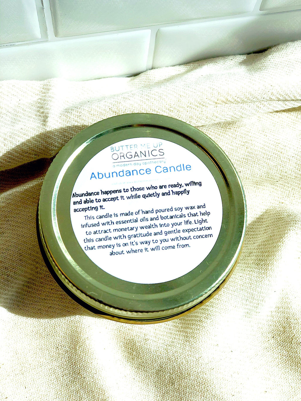 Close-up of the gold lid label on a soy wax abundance candle detailing its manifestation properties, set against a white subway tile kitchen backsplash.