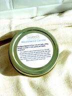 Close-up of the gold lid label on a soy wax abundance candle detailing its manifestation properties, set against a white subway tile kitchen backsplash.